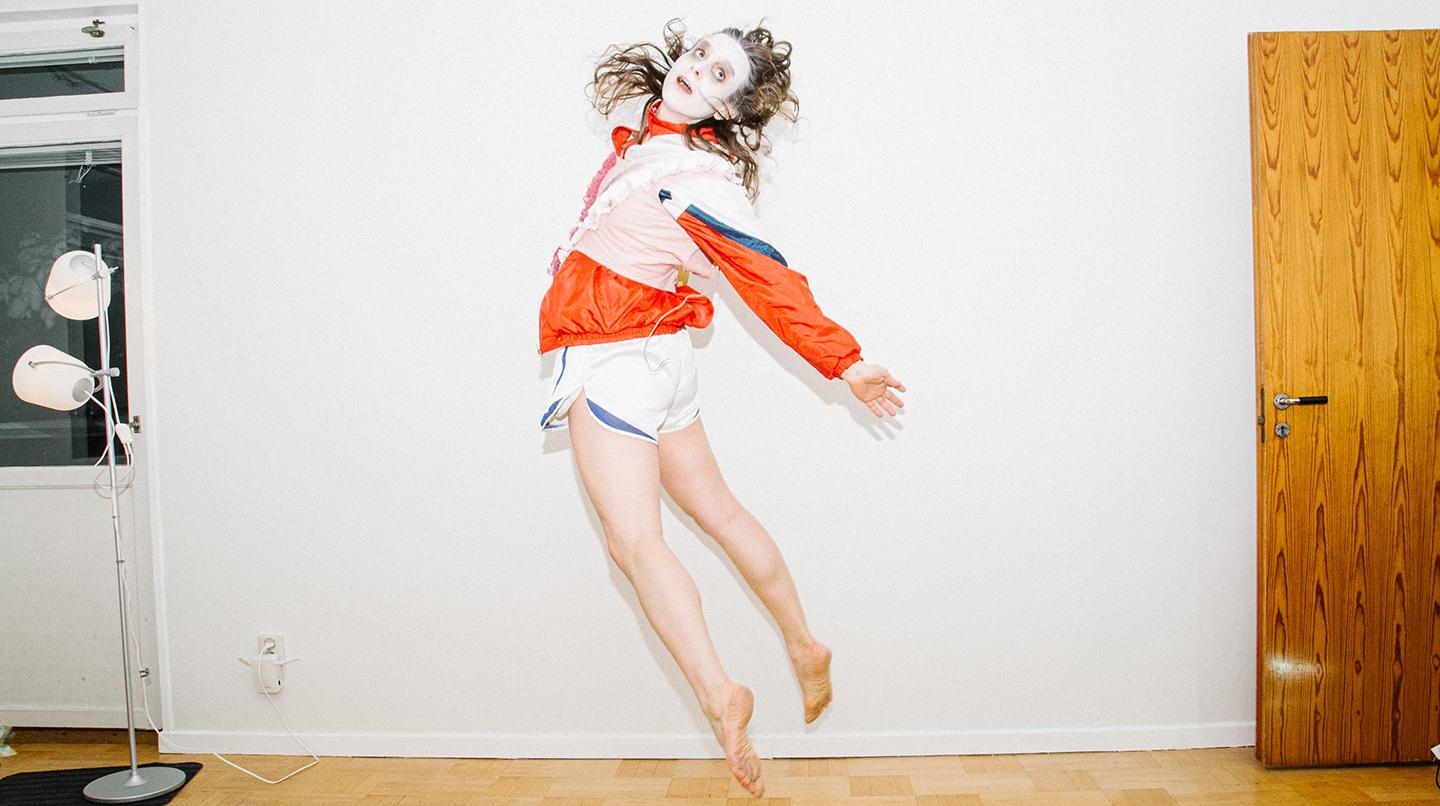 A woman in white make-up, wearing shorts and a windbreaker, jumps into the air and looks over her shoulder