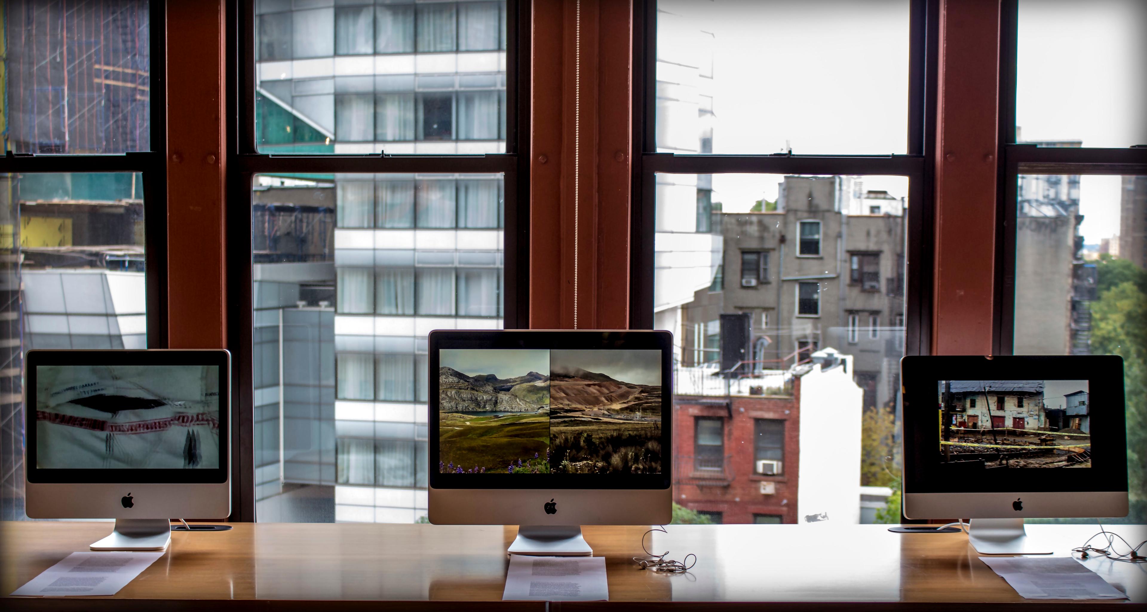 Three Apple computers, each with a piece of paper with text in front of them, stand on a table by a window looking out onto houses and buildings.